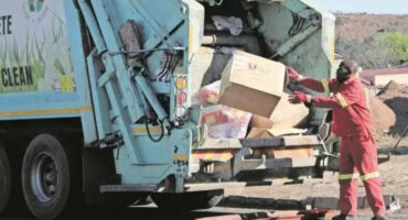 A man in a red shirt is loading trash into a garbage truck on a city street.