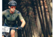 A cyclist in a green jersey and helmet rides intently through a forest. Sunlight filters through the trees, creating a determined and focused mood.