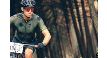 A cyclist in a green jersey and helmet rides intently through a forest. Sunlight filters through the trees, creating a determined and focused mood.