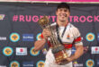 A man proudly holds a trophy in front of a decorative wall, showcasing his achievement.