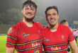 Two rugby players wearing red jerseys smile broadly on a rainy field. Their shirts display logos and their camaraderie conveys joy and achievement.