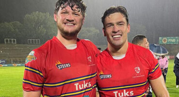 Two rugby players wearing red jerseys smile broadly on a rainy field. Their shirts display logos and their camaraderie conveys joy and achievement.