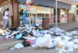 A cluttered street corner with overflowing trash, including bags and discarded items. Two pedestrians navigate the rubbish, creating a chaotic scene.