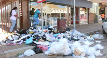 A cluttered street corner with overflowing trash, including bags and discarded items. Two pedestrians navigate the rubbish, creating a chaotic scene.
