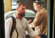 Two men are seen outdoors beside a white car, wearing casual shirts and carrying backpacks. One appears thoughtful, the other focused, suggesting a serious tone.