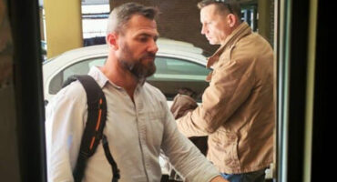 Two men are seen outdoors beside a white car, wearing casual shirts and carrying backpacks. One appears thoughtful, the other focused, suggesting a serious tone.