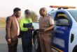 Three men converse by a police vehicle on a road. One in a suit, another in a neon jacket, and the third, a police officer, in uniform holding a tablet.