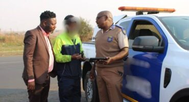 Three men converse by a police vehicle on a road. One in a suit, another in a neon jacket, and the third, a police officer, in uniform holding a tablet.