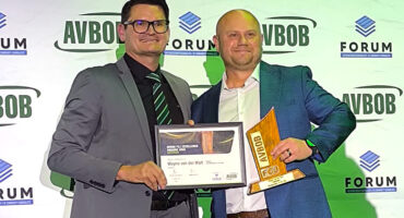 Two men proudly display an award in front of a decorative wall.