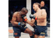Two men engage in a cage fight, with one man on the ground and the other standing above him, ready to strike.