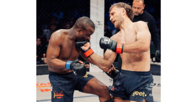Two men engage in a cage fight, with one man on the ground and the other standing above him, ready to strike.