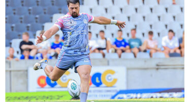 A rugby player in action, preparing to kick the ball on a grassy field during a match.