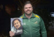 A man holds a book titled "Tana Umaga," showcasing the cover prominently in his hands.