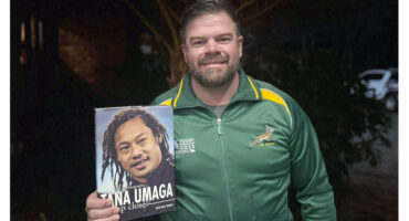 A man holds a book titled "Tana Umaga," showcasing the cover prominently in his hands.