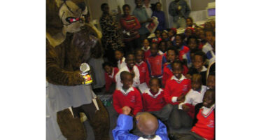 A group of children sitting on the floor in a room, smiling and interacting with a colorful mascot character.