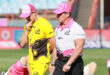 Two men dressed in a yellow and a pink shirt and pink hats pose on a green field, showcasing a friendly demeanor