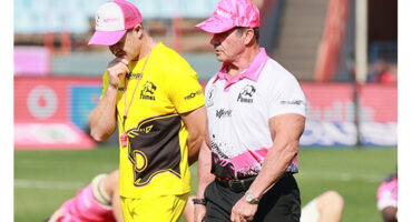 Two men dressed in a yellow and a pink shirt and pink hats pose on a green field, showcasing a friendly demeanor
