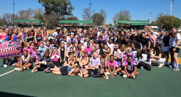 A group of girls smiling and posing together on a netball court, with netball equipment visible in the background.