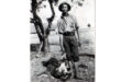 A man stands on grass, holding a crocodile, with a neutral expression and casual attire
