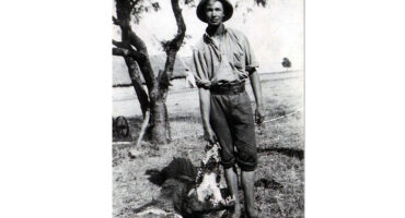 A man stands on grass, holding a crocodile, with a neutral expression and casual attire