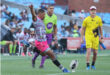 A rugby player in a pink shirt executing a kick towards the ball on a grassy field.