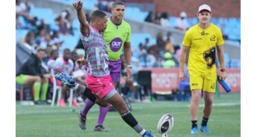 A rugby player in a pink shirt executing a kick towards the ball on a grassy field.