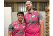 Two men wearing pink and white jerseys stand side by side, smiling at the camera.