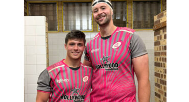 Two men wearing pink and white jerseys stand side by side, smiling at the camera.
