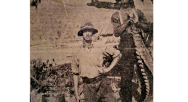 Two men stand beside a large tree in an old photograph, showcasing their attire and the natural surroundings. The one is holding a dead crocodile.