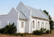 A white church featuring a tin roof is centrally located in a vast green field, surrounded by open space.