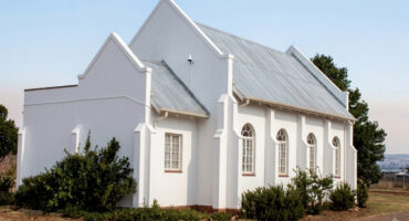 A white church featuring a tin roof is centrally located in a vast green field, surrounded by open space.