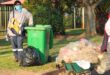 A man wearing a mask and gloves stands beside a garbage can, emphasizing safety and hygiene practices.