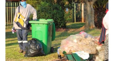 A man wearing a mask and gloves stands beside a garbage can, emphasizing safety and hygiene practices.
