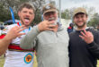 Three men smiling and posing together at a sporting event, wearing team jerseys and surrounded by fans.
