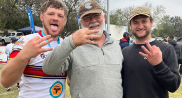 Three men smiling and posing together at a sporting event, wearing team jerseys and surrounded by fans.