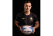 A man in a black shirt stands holding a rugby ball, ready to pass or throw it during a game.