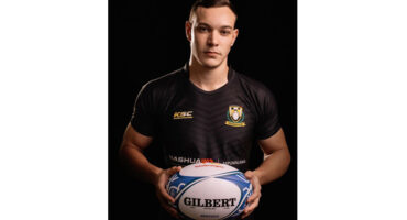 A man in a black shirt stands holding a rugby ball, ready to pass or throw it during a game.
