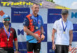 Three men stand on a podium, each wearing medals; one man proudly holds his medal aloft.