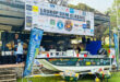 A boat is docked in front of a tent displaying a sign, indicating the location or event taking place.