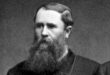 Black and white photo of a man with a long beards, looking directly at the camera with a serious expression.