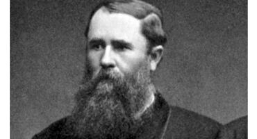 Black and white photo of a man with a long beards, looking directly at the camera with a serious expression.