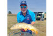 A man proudly holds a large fish in a grassy field, showcasing his catch of the day.