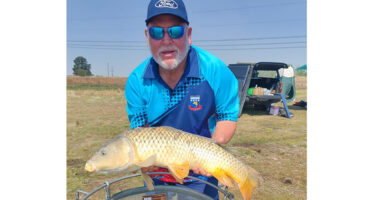 A man proudly holds a large fish in a grassy field, showcasing his catch of the day.