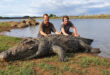 Two men stand on the shore, smiling beside a large crocodile resting on the sand.
