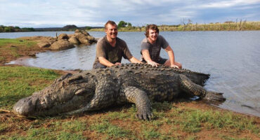Two men stand on the shore, smiling beside a large crocodile resting on the sand.