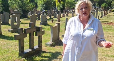 A woman gazes thoughtfully at a cross in a cemetery, surrounded by gravestones, evoking a sense of loss and remembrance.
