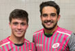 Two young men stand side by side, one in a pink shirt and the other in a black shirt, smiling at the camera.