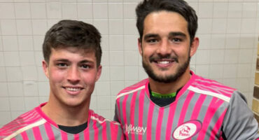 Two young men stand side by side, one in a pink shirt and the other in a black shirt, smiling at the camera.