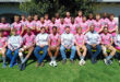 A diverse group of men and women wearing matching pink shirts, smiling and standing together outdoors.