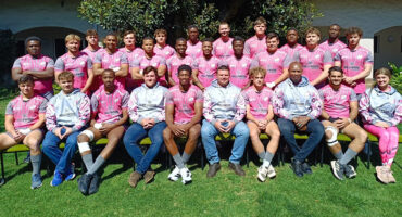 A diverse group of men and women wearing matching pink shirts, smiling and standing together outdoors.